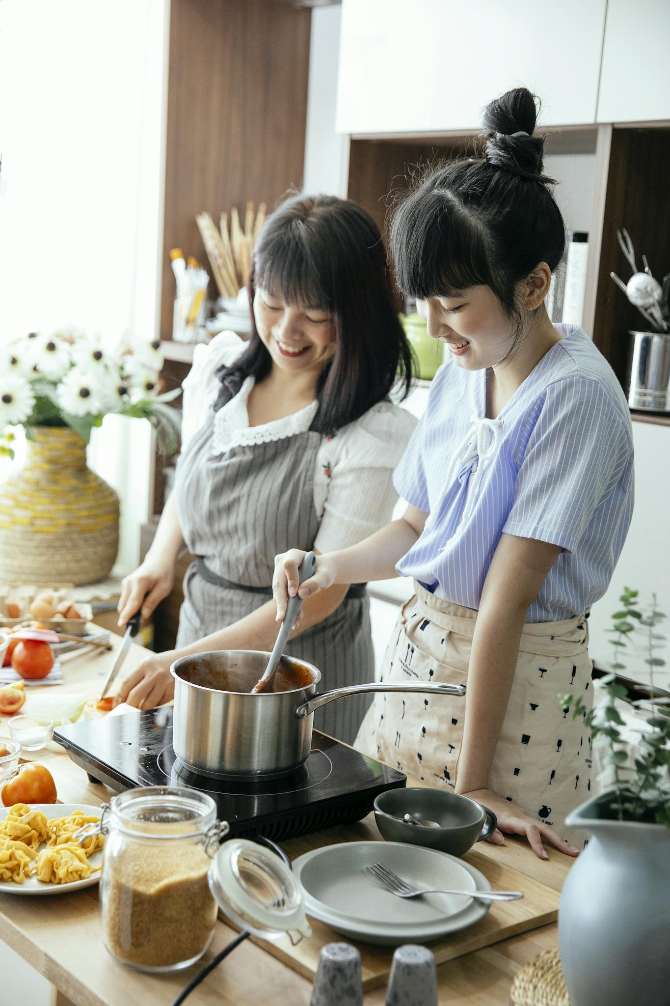 Cheerful Asian woman with young daughter smiling while cutting veggies and stirring sauce during lunch preparation together in kitchen